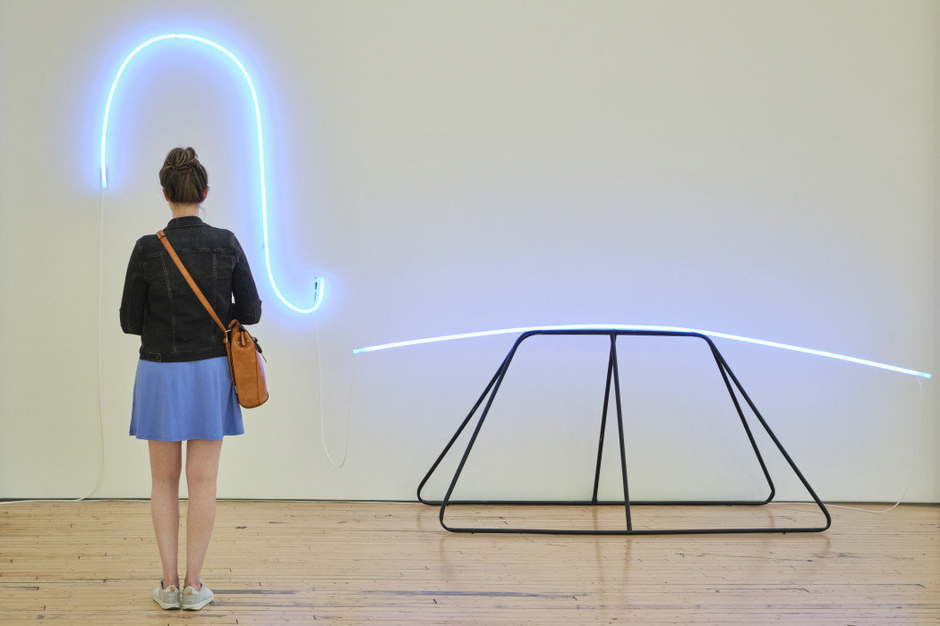 Woman stands in front of a sculpture made of metal and blue neon. The neon loops on the wall and frames the woman.